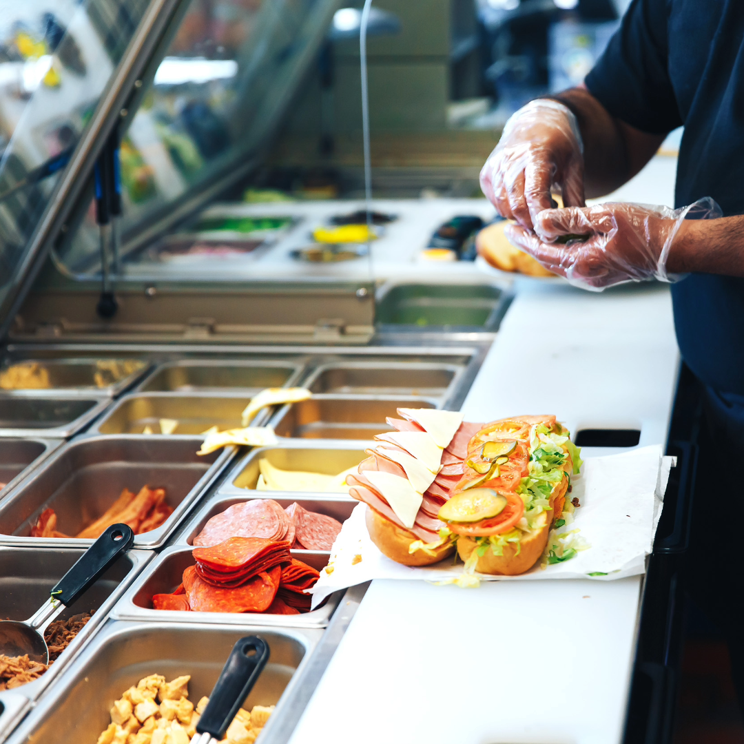 QSR employee wearing gloves prepping a sandwich at the food prep station.