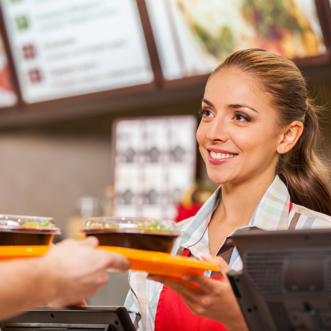 A woman in an apron behind the register handing someone a tray of food.