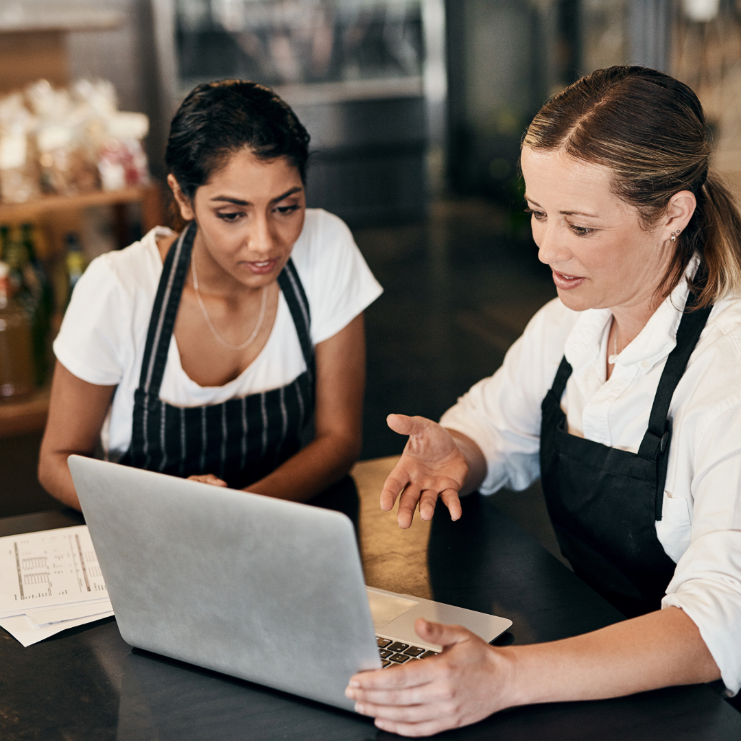 Two women in aprons sitting at a table, looking at a laptop and talking.