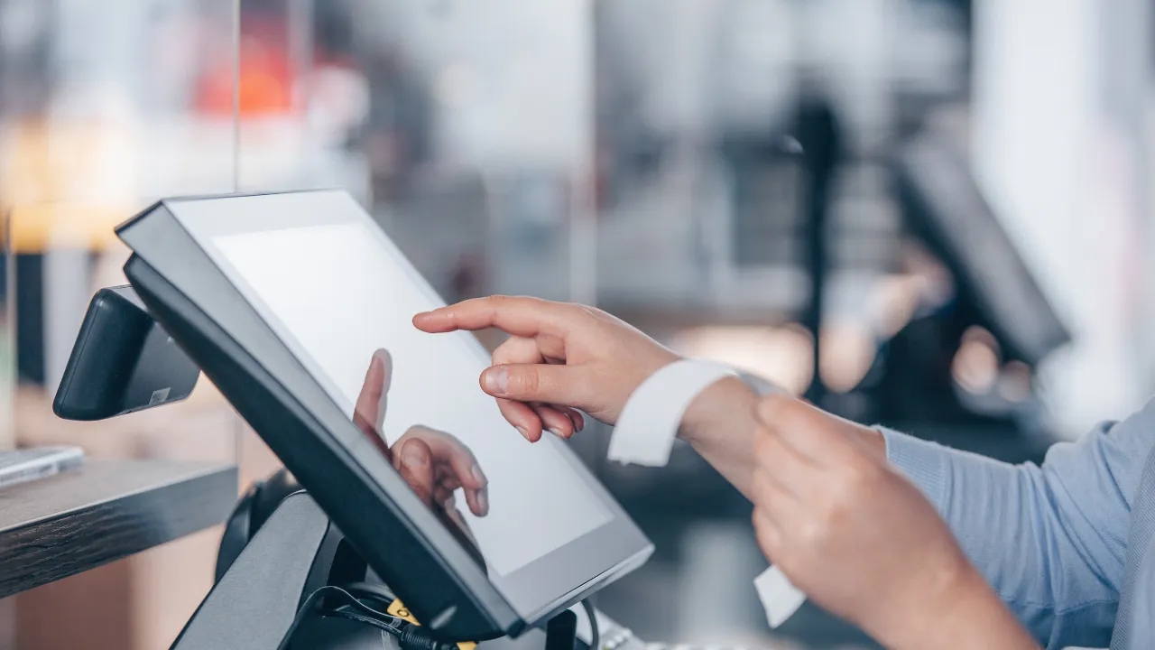Close-up of a store employee tapping a touchscreen POS terminal while holding a receipt, highlighting where manual inputs trigger POS data discrepancies.