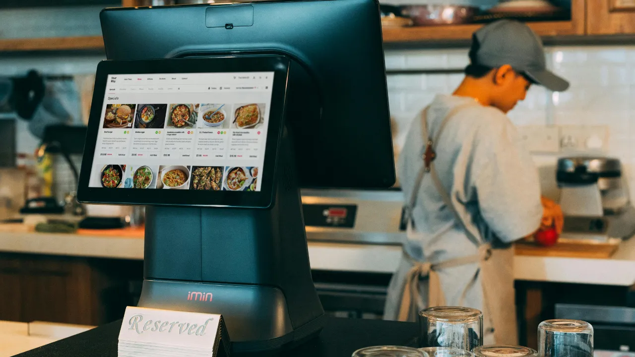 Touchscreen point of sale ordering terminal on a restaurant counter with a staff member working in the background.
